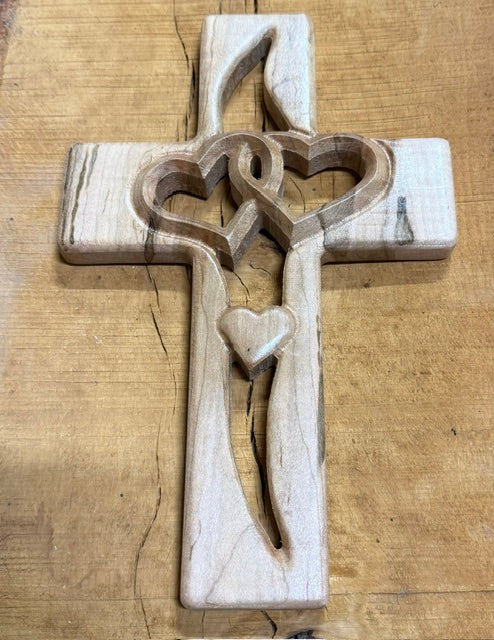 A carved wooden cross with a central heart-shaped cutout and a small heart near the bottom, displayed on a rustic wooden surface.