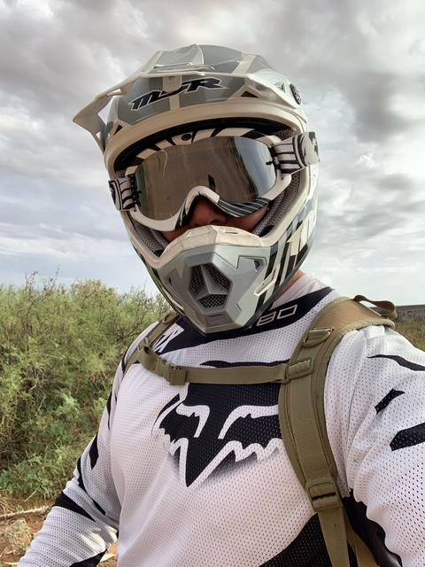A person outdoors wearing a white and black Fox Racing motocross jersey, a matching helmet with a clear visor, and a tan backpack against a cloudy sky and grassy background. model  for carving 