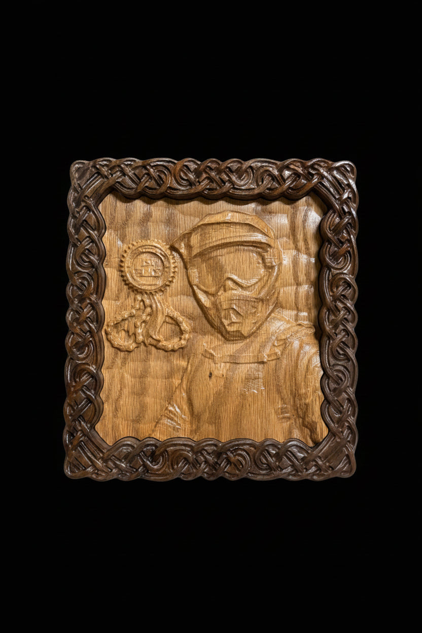 A square wooden carving plaque featuring a relief portrait of a person in a helmet and goggles, holding a ribbon medallion, surrounded by an ornate braided knot border. The plaque is photographed on a light fabric surface with a wood grain background.